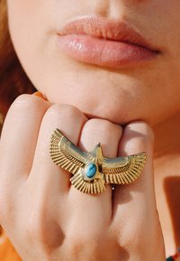 Bague en or en forme d'aigle avec des détails de plumes complexes et une pierre turquoise au centre, portée sur une main avec une peau légèrement texturée.