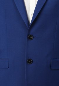 Blå blazer med to sorte knapper, slående lapelkraver og et tekstureret stof. Inkluderer to forlommer og en hvid skjorte under.