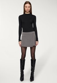 Czarna bluzka z długim rękawem w połączeniu z szarym owijanym spódnicą z przednią kieszenią. Modelka ma na sobie czarne kozaki za kolano na obcasie o błyszczącej powierzchni.