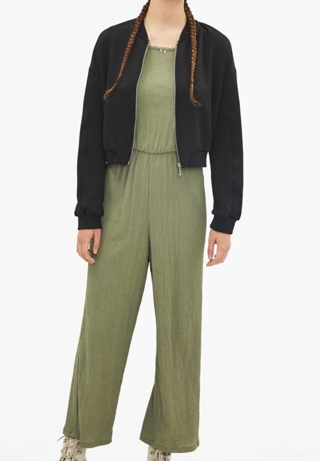 Personne portant une veste zippée noire sur une combinaison verte texturée avec jambes larges et des chaussures claires.