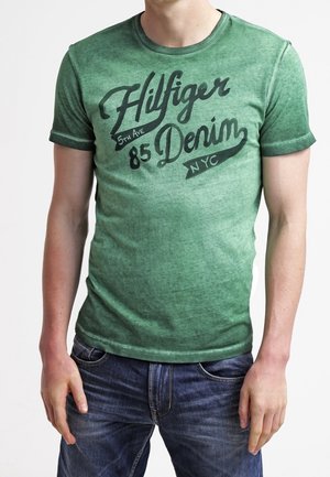 Uomo che indossa una maglietta verde Hilfiger Denim con scritte nere e jeans blu, in piedi con le mani vicino alle tasche.