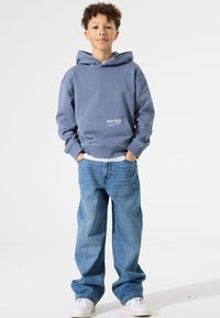Blauer Hoodie mit Kängurutasche und weißem Textdetail, kombiniert mit locker sitzenden hellblauen Jeans und weißen Sneakers. Glatte Stoffstruktur.