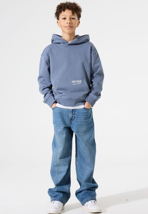 Blauwe hoodie met een kangoeroezak en witte tekstdetails, gecombineerd met losse lichtblauwe jeans en witte sneakers. Gladde stoftextuur.