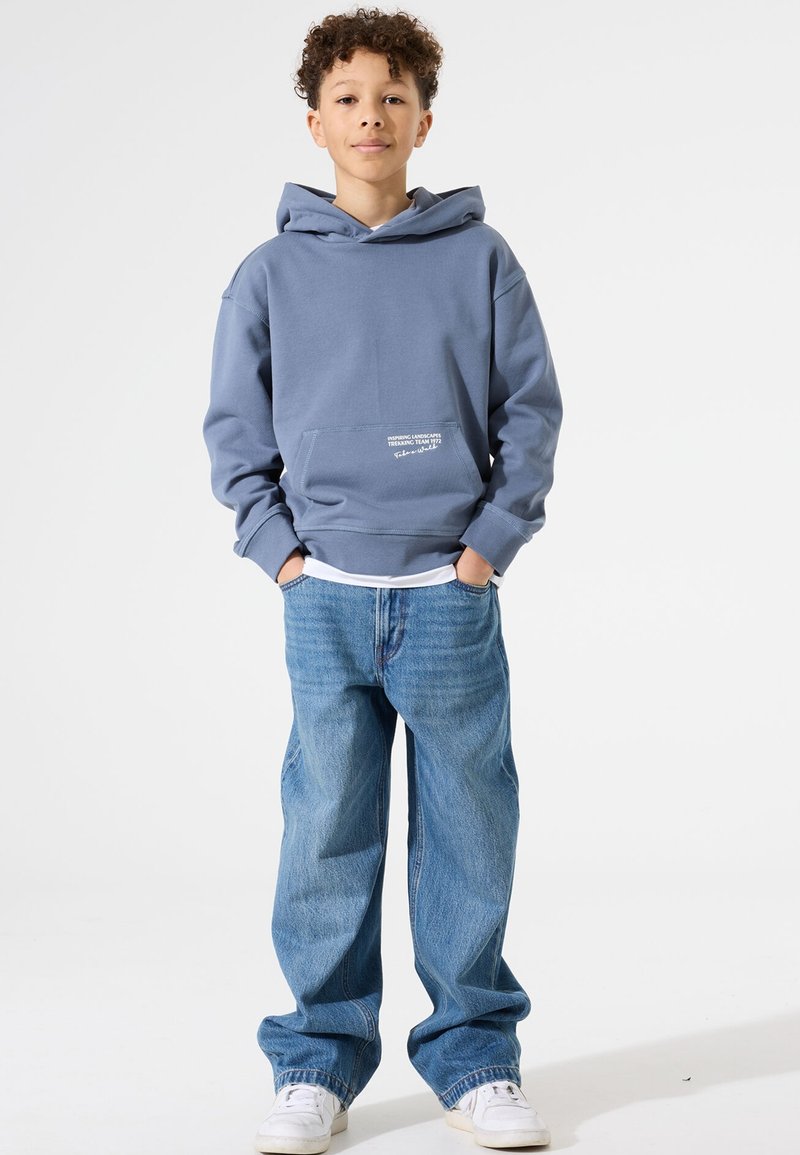 Blauer Hoodie mit Kängurutasche und weißem Textdetail, kombiniert mit locker sitzenden hellblauen Jeans und weißen Sneakers. Glatte Stoffstruktur.