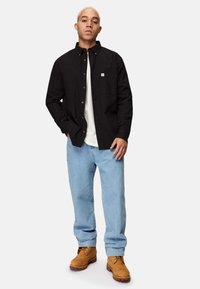 Sort skjorte med knapper og en forlomme, parret med lyseblå løstsiddende jeans og brune snørebåndsstøvler, stående mod en ensfarvet baggrund.