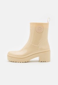 Beige rubberen enkellaars met een ronde neus, dikke hak en een ronde logo-accent aan de zijkant. Heeft een gladde, glanzende afwerking.