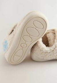 Chaussons beiges doux avec une texture en peluche ; la semelle en caoutchouc présente des motifs de grippe ovales et une indication de taille 6 ; accent brodé bleu.