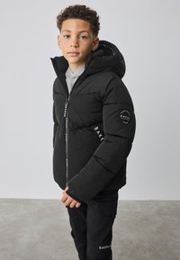 Zwarte pufferjas met capuchon, ritssluiting aan de voorkant en merkdetails. Heeft een doorgestikte textuur en manchetten aan de mouwen.