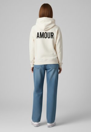 Felpa crema con la scritta "AMOUR" stampata in nero sulla schiena, abbinata a jeans leggeri blu con motivo e sneakers bianche con accenti rossi.