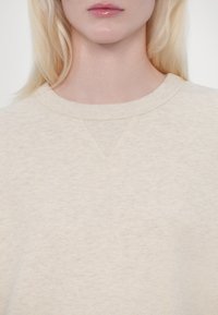 Personne blonde aux lèvres brillantes portant un pull beige clair à col rond avec un détail en V côtelé et cousu au niveau du col.