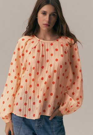 Woman wearing a loose, light peach blouse with red polka dots and long puffed sleeves, paired with blue denim pants, neutral background.