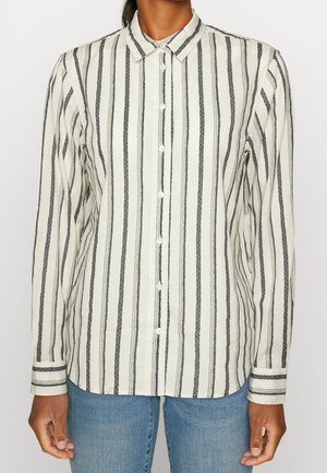 Personne portant une chemise blanche boutonnée avec des motifs à rayures noires verticales et un jean bleu, debout devant un fond uni.