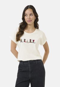 T-shirt en coton de couleur crème présentant une rangée de bottes illustrées dans différents styles et couleurs. Porté avec un pantalon en denim noir.