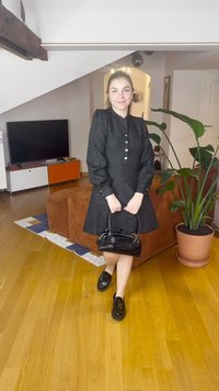 Melnā kleita ar pūstām piedurknēm, aizdare ar pogām, teksturēts raksts; kombinēta ar melnu rokassomu un loaferiem, iekštelpās ar mājīgu atmosfēru.