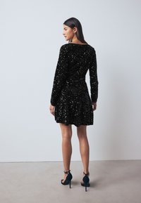 Robe noire en sequins avec manches longues, corsage ajusté, jupe évasée, découpe au dos, associée à des sandales à talons hauts noires.