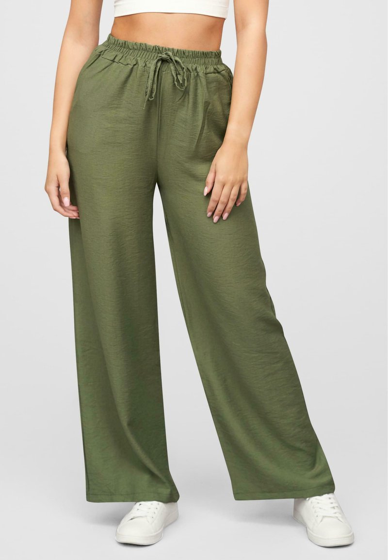 Pantalones de pierna ancha en verde oliva con cinturilla elástica y cordón, confeccionados en una tela suave y ligera. Combinados con zapatillas blancas.