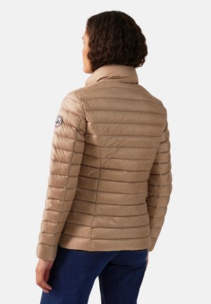 Mujer con cabello rizado corto, vistiendo una chaqueta acolchada beige y jeans azul oscuro, vista desde atrás contra un fondo liso.