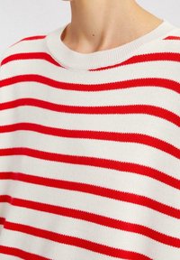 Nahaufnahme einer Person, die einen weißen Pullover mit horizontalen roten Streifen und einem gerippten Rundhalsausschnitt trägt.