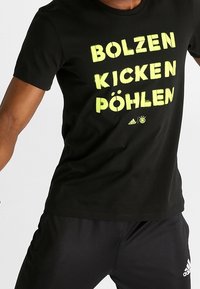Camiseta negra con texto amarillos en negrita "BOLZEN KICKEN PÖHLEN." Hecha de una tela suave con cuello redondo y mangas cortas.