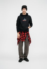 Crna dukserica s šarenim grafičkim uzorkom, crvena i crna karo košulja zavezana oko struka, sivi hlače i crne tenisice.