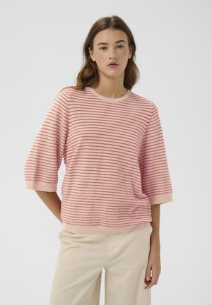 Femme portant un pull à manches courtes rayé rose et rouge et un pantalon beige clair, debout devant un fond clair uni.