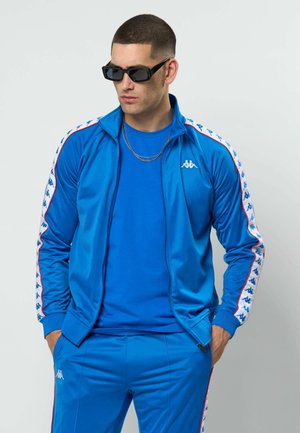 Férfi, kék Kappa melegítőben, fehér logósávokkal, fekete napszemüvegben, zsebébe dugott kézzel, oldalra nézve.