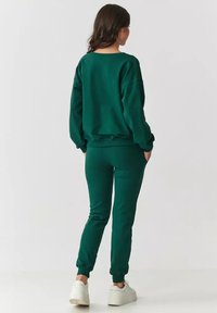 Zielony bawełniany sweter z długim rękawem i dopasowane dresy z prążkowanymi mankietami, w połączeniu z białymi sneakersami. Prosty design, miękka tekstura i luźny krój.
