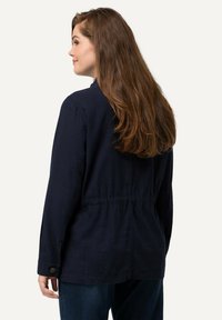 Marineblauwe, lange mouw jacket gemaakt van een gestructureerde stof, met een aangesloten taille en knoopmanchetten. De achterkant toont een ontspannen pasvorm ontwerp.