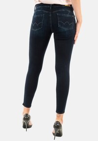 Jeans en denim noir skinny avec une taille mi-haute, dotés de poches arrière courbes et d'une texture lisse. Portés avec des chaussures à talons hauts noires.