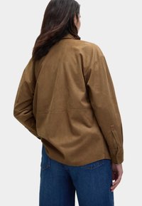 Chemise en daim marron avec une coupe décontractée, épaules basses et manches longues. Présente un détail de couture à l'arrière et est associée à un jean en denim bleu.