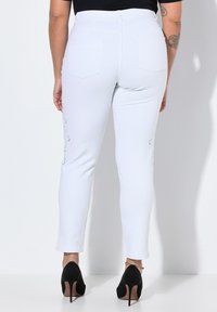 Jeans bianchi aderenti con una texture liscia, caratterizzati da ricami decorativi sui lati e nelle tasche posteriori, abbinati a scarpe nere con tacco alto.