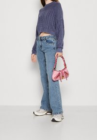 Sweter w stylu szydełkowym w kolorze fioletowym, jasnoniebieskie dżinsy z rozszerzanymi nogawkami oraz mała różowa torebka z złotymi akcentami, noszona z szaro-białymi trampkami.