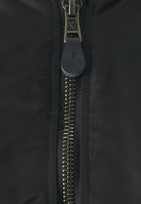 Svart jakke med en sentral metallglidelås med messingtenner og en lærpull-tab, glatt nylonstoff og uten distinkte mønstre.
