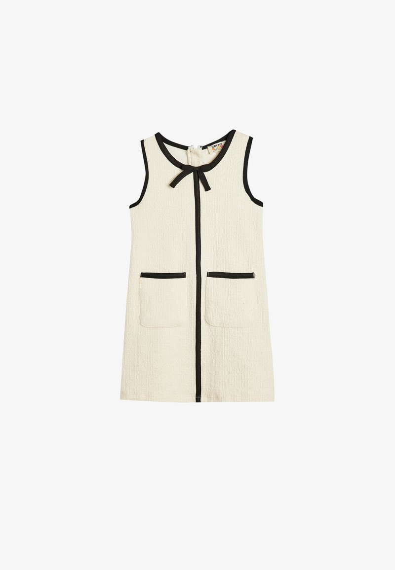 Kremowa sukienka bez rękawów z teksturowanej tkaniny, czarny wykończenia wzdłuż krawędzi, dwie kieszenie z przodu oraz detal w postaci kokardy przy dekolcie.