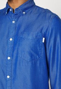 Homme portant une chemise en jean bleue à boutons avec une poche poitrine et des boutons blancs, col boutonné.