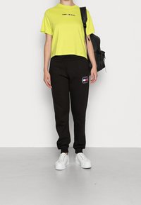Lime vihreä lyhythihainen t-paita, jossa on "TOMMY JEANS" -logo; mustat jogger-housut, joissa on logopatch; valkoiset lenkkarit; musta reppu.