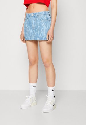 Kobieta w niebieskiej mini spódniczce denimowej z wzorami, czerwonym krótkim topie, białych skarpetkach Nike i białych tenisówkach Nike na jednolitym tle.