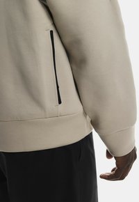 Hanorac bej cu buzunar lateral cu fermoar, textură moale și design ajustat. Mansetele și tivul sunt cu nervuri, creând un aspect elegant.