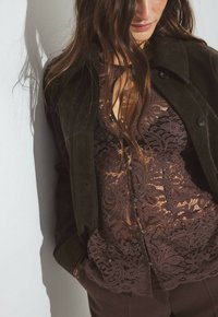 Top in pizzo marrone scuro sovrapposto a una giacca in suede verde scuro, con chiusura a bottone e intricati motivi floreali. Tessuto morbido ben visibile.