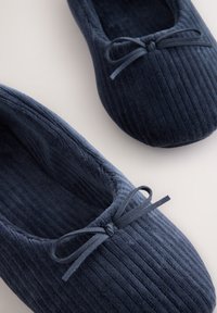 Paire de chaussons en velours côtelé bleu marine avec petits nœuds noués sur le dessus, montrée sur une surface claire et unie.