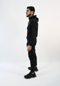 Chaqueta atlética negra con capucha, combinada con pantalones negros y zapatillas. La tela parece ligera; un logo es visible en la chaqueta.