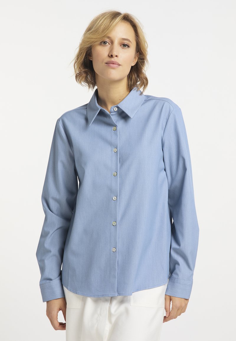 Woman with short blonde hair wearing a light blue button-up shirt and white pants against a plain white background.