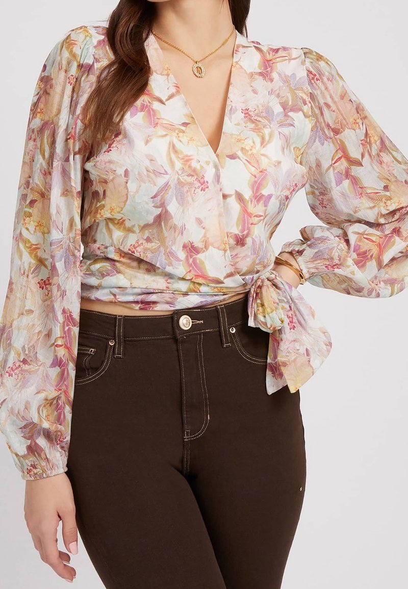 Haut cache-cœur floral en tissu transparent, avec de longues manches bouffantes, un lien à la taille, et un mélange de couleurs pastel avec des motifs de feuilles. Associé à un jean marron foncé.