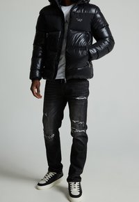 Schwarze Pufferjacke mit Reißverschluss, gepolstertem Design und kleinem Logo. Trägt sie zu ausgefransten schwarzen Jeans und schwarz-weißen Sneakers.