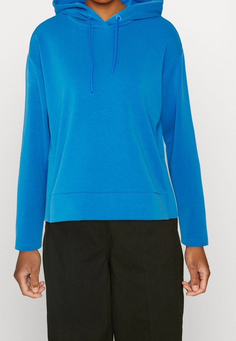 Modrá mikina s kapucí a dlouhými rukávy, vyrobená z měkkého materiálu. Má stahovací šňůrky a mírně zkrácený lem. Kombinována s tmavými kalhotami.