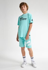 Camiseta de color verde claro con el logotipo "Gran Turismo" en negro, combinada con pantalones cortos a juego que presentan acentos de logo en blanco. Completado con zapatillas blancas y calcetines a rayas.