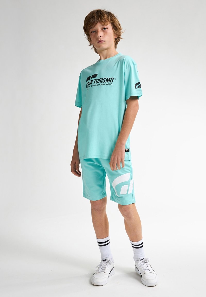 Camiseta de color verde claro con el logotipo "Gran Turismo" en negro, combinada con pantalones cortos a juego que presentan acentos de logo en blanco. Completado con zapatillas blancas y calcetines a rayas.