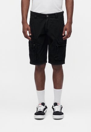 Uomo con pantaloncini cargo neri, calzini bianchi classici e scarpe da ginnastica bianche e nere, in piedi su sfondo bianco neutro.