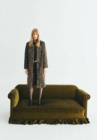 Groene fluwelen bank met franje; een vrouw staat erop, gekleed in een leopardprintjas en zwarte kleding, met lang haar.