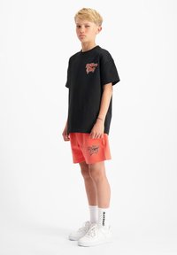 Zwart T-shirt met korte mouwen en rode logo, gecombineerd met koraalkleurige shorts met bijpassend logo; witte sneakers en crew-length sokken.